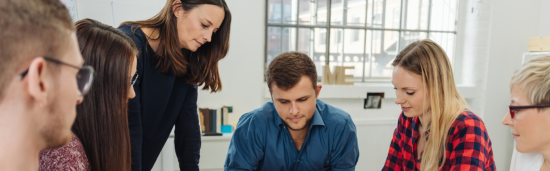 Zu sehen ist eine Gruppe junger Menschen. Sie sind in der Ausbildung. Gemeinsam sitzen sie um einen Tisch herum. Die Ausbilderin steht und erklärt den jungen Menschen etwas. Sie hören alles zu.