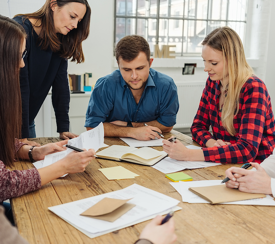 Zu sehen ist eine Gruppe junger Menschen. Sie sind in der Ausbildung. Gemeinsam sitzen sie um einen Tisch herum. Die Ausbilderin steht und erklärt den jungen Menschen etwas. Sie hören alles zu.