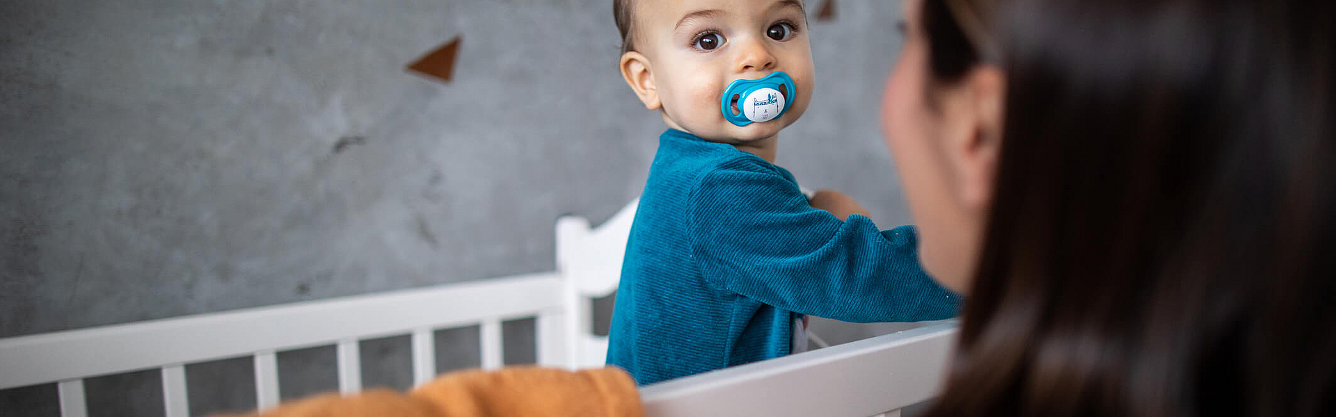Kleinkind steht im Kinderbett, die Mama lächelt es an
