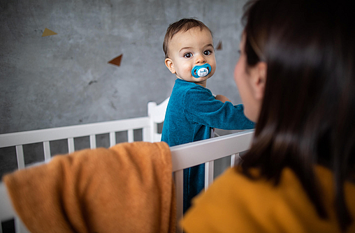 Kleinkind steht im Kinderbett, die Mama lächelt es an