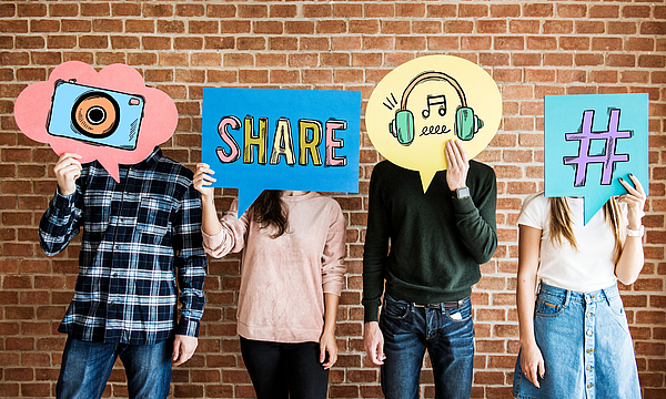 Eine Gruppe steht vor einer Mauer. Sie halten Plakate mit Social-Media-Icons hoch. Zum Beispiel den Hashtag.