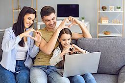 Familie sitzt auf einem Sofa. Sie videochatten und formen die Hände zu Herzen.
