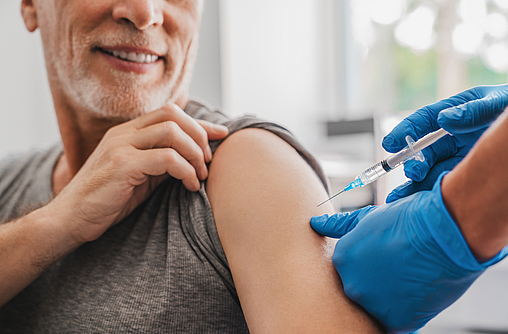 Ein Mann im mittleren Alter mit Bart hat den linken Ärmel seines Shirts hochgekrämpelt. Er bekommt mit einer Spritze eine Impfung. Die Impfung macht ein Arzt.