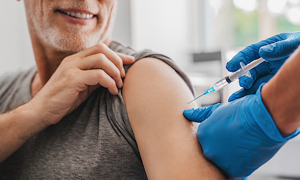Ein Mann im mittleren Alter mit Bart hat den linken Ärmel seines Shirts hochgekrämpelt. Er bekommt mit einer Spritze eine Impfung. Die Impfung macht ein Arzt.