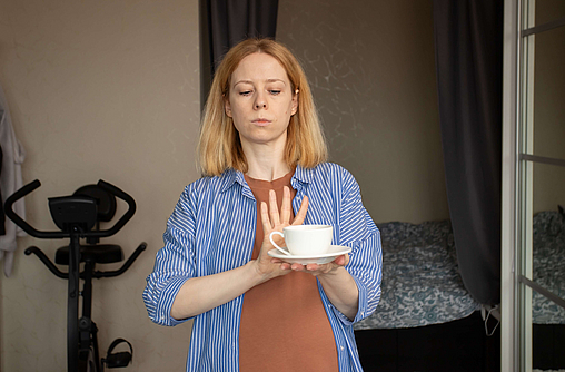 Schwangere Frau hält eine Tasse Kaffee in der Hand und macht mit der anderen Hand eine streckt die andere Hand der Tasse ablehnend entgegen.
