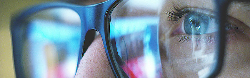 Nahaufnahme eines Mannes. Er trägt eine schwarze Brille und schaut auf einem Monitor. Dieser spiegelt sich im Brillenglas.