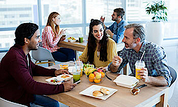 Drei Kollegen sitzen zusammen an einem Holztisch. Sie essen und unterhalten sich. Im Hintergrund sind zwei weitere Personen, die sich unterhalten und währenddessen essen. Alle befinden sich in einem Büro.