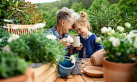 Oma sitzt mit ihrer kleinen Enkeltochter auf der Terrasse im Grünen und sind glücklich. Auf dem Tisch stehen verschiedene Pflanzentöpfe.