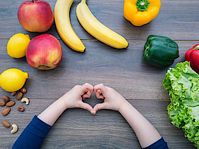 Auf einem steinernen Tisch liegt verschiedenes Obst und Gemüse: Bananen, Nüsse, Paprikas, eine Zitrone und ein Apfel und ein Salat. Ein Kind bildet mit seinen Fingern nein Herz.