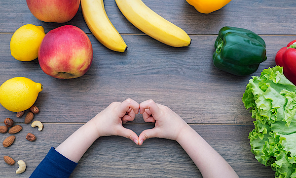 Auf einem steinernen Tisch liegt verschiedenes Obst und Gemüse: Bananen, Nüsse, Paprikas, eine Zitrone und ein Apfel und ein Salat. Ein Kind bildet mit seinen Fingern nein Herz.