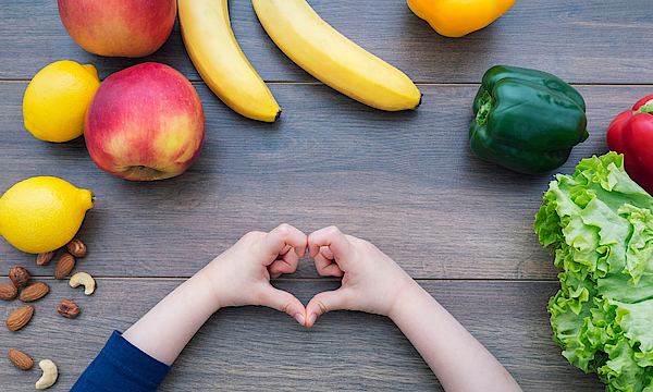 Auf einem steinernen Tisch liegt verschiedenes Obst und Gemüse: Bananen, Nüsse, Paprikas, eine Zitrone und ein Apfel und ein Salat. Ein Kind bildet mit seinen Fingern nein Herz.