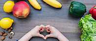 Auf einem steinernen Tisch liegt verschiedenes Obst und Gemüse: Bananen, Nüsse, Paprikas, eine Zitrone und ein Apfel und ein Salat. Ein Kind bildet mit seinen Fingern nein Herz.