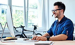 Ein Mann sitzt an einem Holztisch. Er trägt ein Headset und arbeitet am Computer