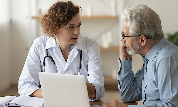 Eine Ärztin in einem weißen Kittel berät einen älteren Patienten. Vor den beiden steht auf einem Tisch ein aufgeklappter Laptop.