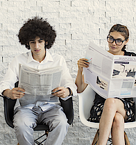 Eine Gruppe junger Menschen sitzt auf Stühlen nebeneinander. Sie lesen alle eine Zeitung.