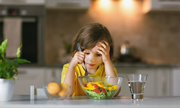 Ein Kind sitzt vor einer Schüssel Gemüse. Daneben steht ein Glas Wasser. Das Kind schaut gelangweilt und verzweifelt.