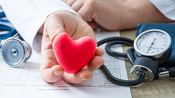 Arzt sitzt am Schreibtisch und hält ein rotes Herz in der Hand.