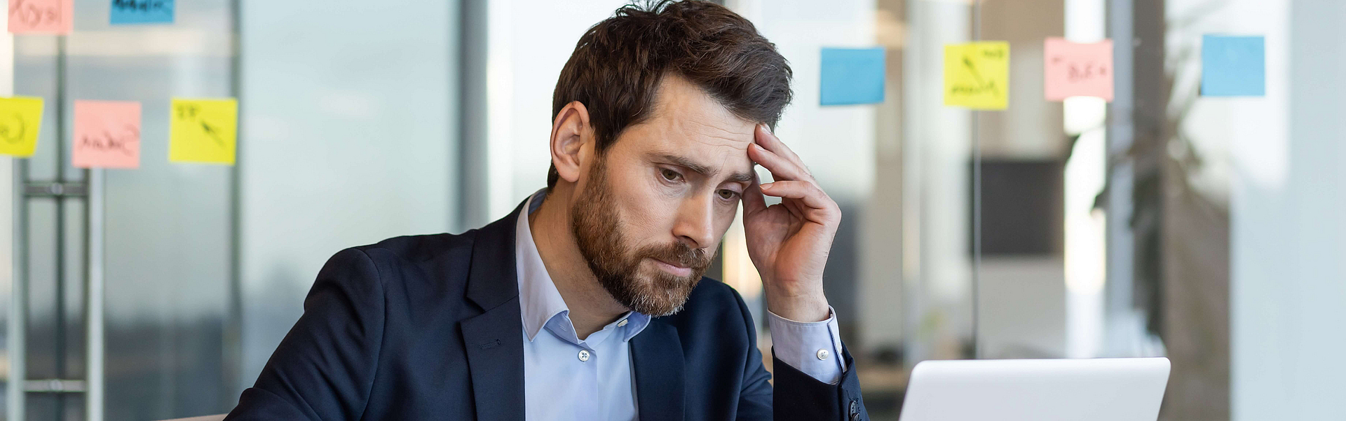 Mann im Anzug sitze am Arbeitsplatz vor einem Laptop, das Gesicht wirkt gestresst
