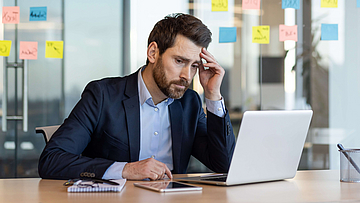 Mann im Anzug sitze am Arbeitsplatz vor einem Laptop, das Gesicht wirkt gestresst