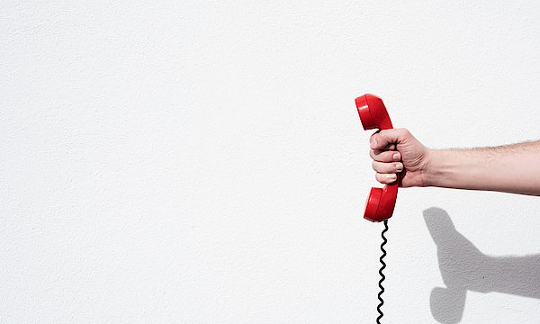 Eine Hand hält einen roten Telefonhörer. An diesem ist eine Telefonschnur befestigt.