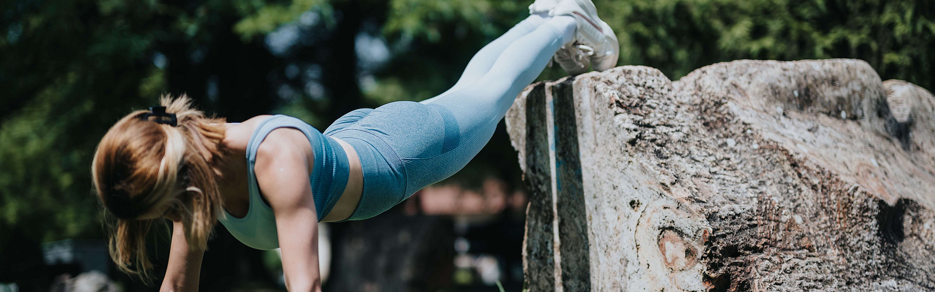 Frau mach eine Liegestütz in einem Park und stellt die Füße dabei auf einen großen Stein. Dieses Krafttraining sorgt für Muskelaufbau und regst so den Stoffwechsel an.