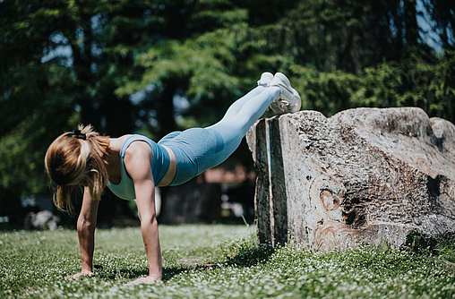 Frau mach eine Liegestütz in einem Park und stellt die Füße dabei auf einen großen Stein. Dieses Krafttraining sorgt für Muskelaufbau und regst so den Stoffwechsel an.
