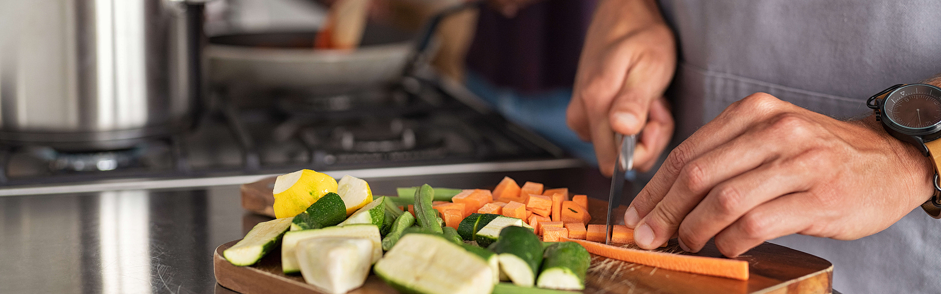 Nahaufnahme von Händen, die Karotten auf dem Scherbrett schneiden. Nahaufnahme von Händen, die Gemüse in der Küche in der Nähe der Brenner schneiden. Detail des Menschen mit Schürze hacken Gemüse für ein Rezept zu Hause.