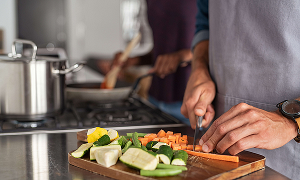 Nahaufnahme von Händen, die Karotten auf dem Scherbrett schneiden. Nahaufnahme von Händen, die Gemüse in der Küche in der Nähe der Brenner schneiden. Detail des Menschen mit Schürze hacken Gemüse für ein Rezept zu Hause.