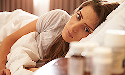 Eine Frau liegt im Bett. Sie schaut auf die Medikamente, die auf dem Nachttisch stehen.