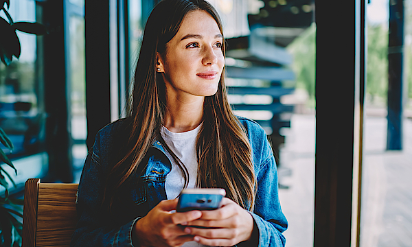 Junge Frau sitzt auf einer Bank und schaut nachdenklich in die Ferne. Das Smartphone hält sie in beiden Händen.