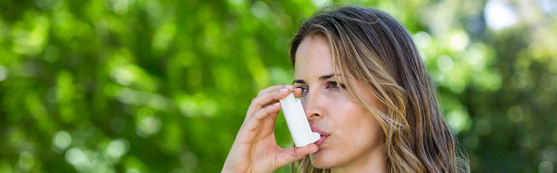 Eine Frau im mittleren Alter ist in der Natur unterwegs. Sie hat blonde Haare mit Locken und ein blau-weiß geringeltes Shirt an. Sie hat Asthma und benutzt ein Asthmaspray.