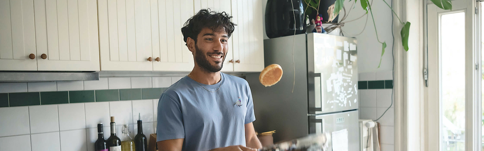 Mann steht in der Küche und wendet einen Pfannkuchen in der Pfanne. Der Pfannkuchen fliegt in die Luft.