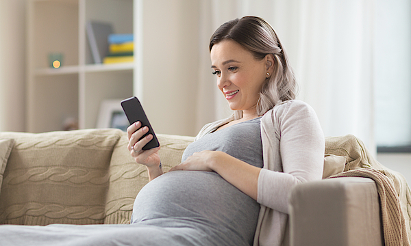 Schwangere sitzt auf dem Sofa und ist mit ihrem Smartphone beschäftigt.