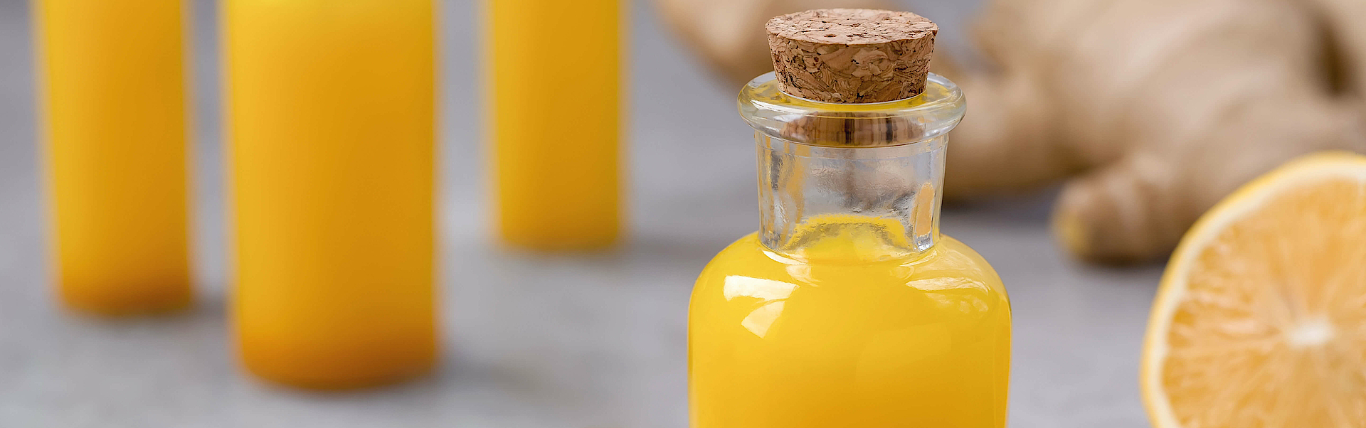 Auf einem Tisch steht ein Ingwer-Kurkuma-Shot in einem Glas. Drum her um sind die Inhaltsstoffe dekoriert: Ingwer, Kurkuma und eine Organge.