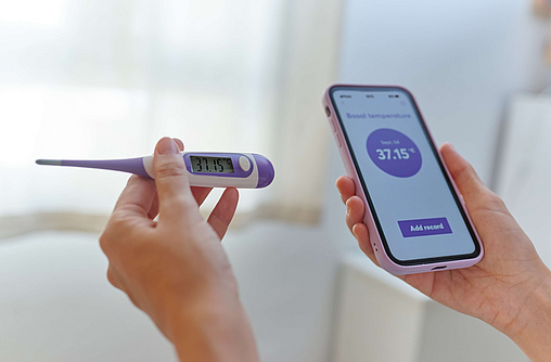 Nahaufnahme von Händen einer Frau, die ein Basalthermometer und Smartphone halten, dass die Basaltemperatur anzeigt.