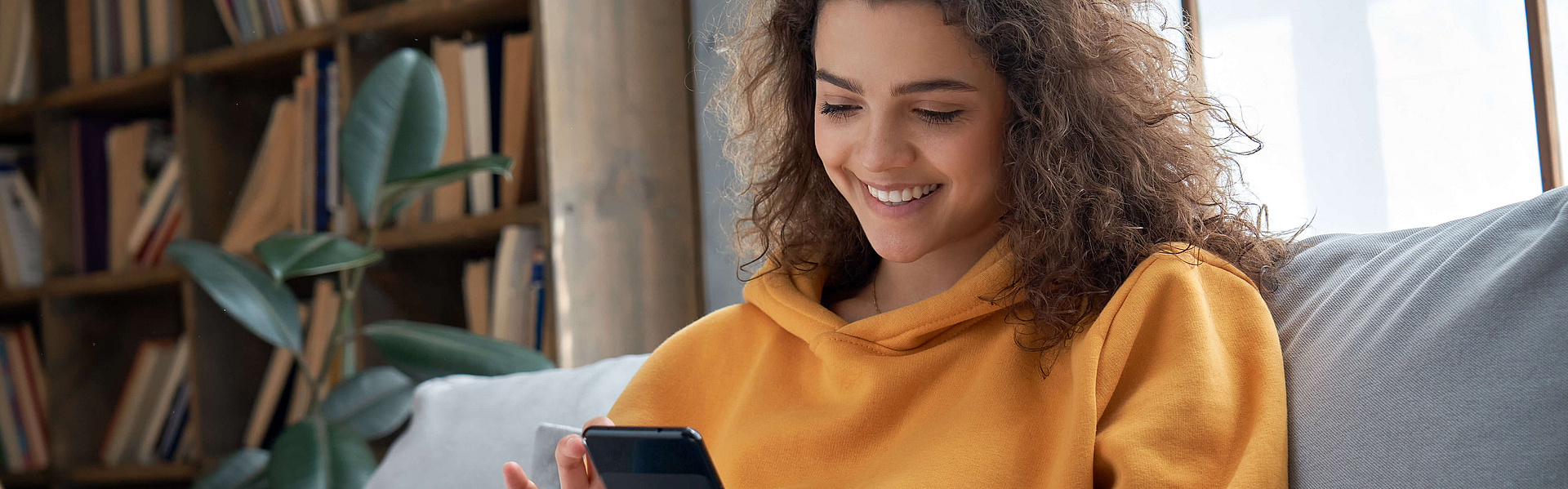 Lächelnd junge Frau sitzt auf dem Sofa und schaut auf ihr Smartphone.