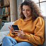 Lächelnd junge Frau sitzt auf dem Sofa und schaut auf ihr Smartphone.