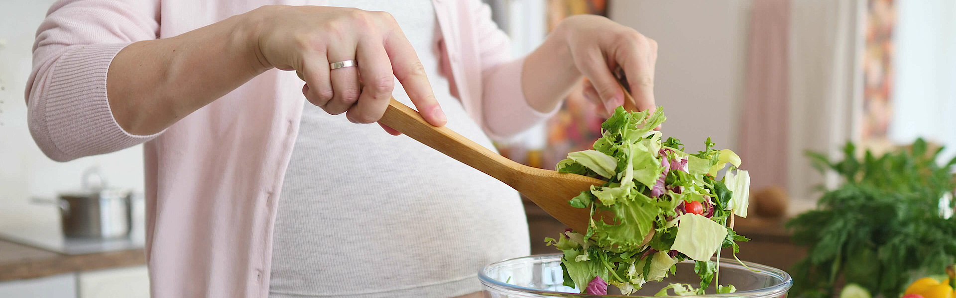 Eine schwangere Frau steht am Küchentisch. Sie macht einen Salat an. 