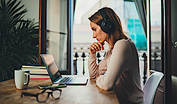 Eine Frau sitzt an einem Tisch und schaut auf den Laptop. Sie trägt ein Headset. Auf dem Tisch liegen Bücher, eine Brille und eine Tasse. Das Fenster im Hintergrund ist geöffnet.