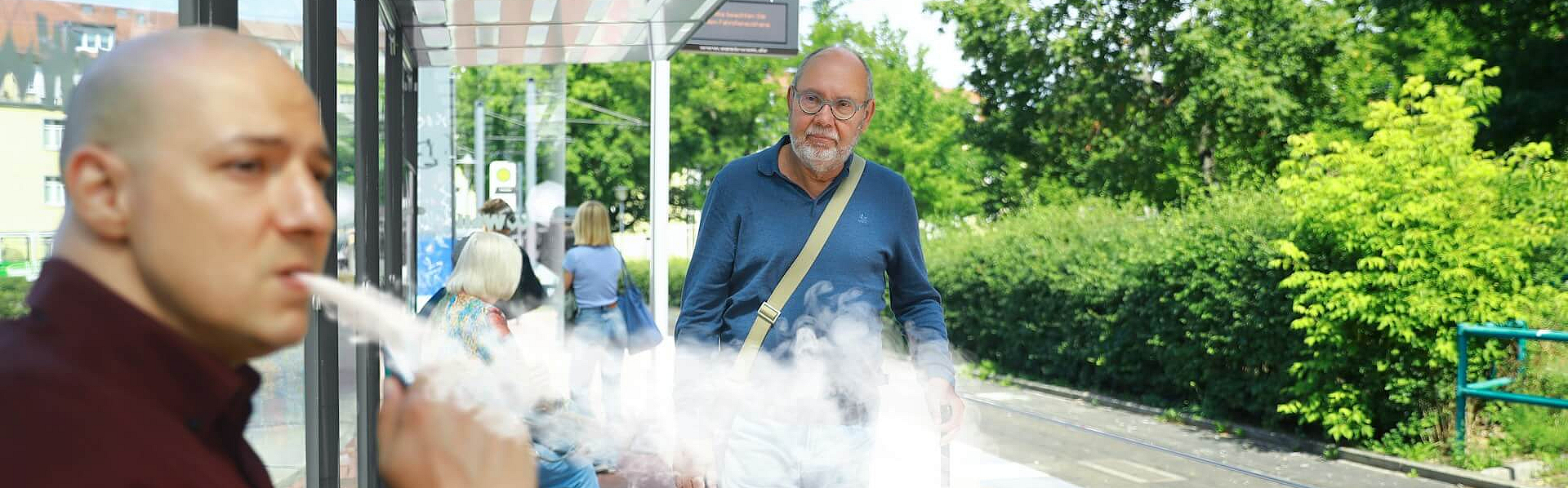 Älterer Mann mit Stock läuft an Bahnsteig auf jungen Mann zu, der dort eine E-Zigarette dampft