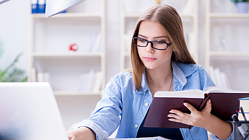 Junge Frau sitzt vor einem Laptop mit einem Buch in der Hand.