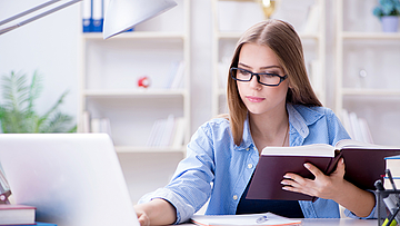 Junge Frau sitzt vor einem Laptop mit einem Buch in der Hand.