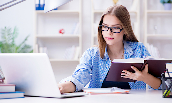Eine junge Frau sitzt vor einem Laptop. Sie hat ein Buch in der Hand und macht sich Notizen. Sie lernt.