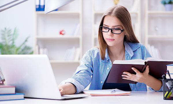 Eine junge Frau sitzt vor einem Laptop. Sie hat ein Buch in der Hand und macht sich Notizen. Sie lernt.