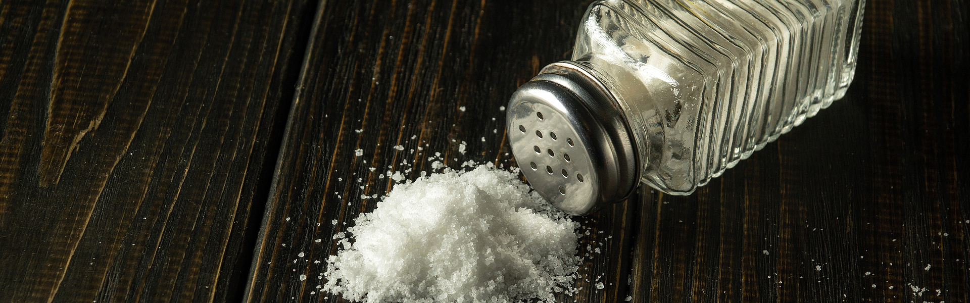 Salzstreuer liegt umgestürzt auf einem dunklen Holz, etwas von dem weißen Salz liegt auf dem Holz