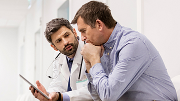 Ein Mann und sein Arzt sitzen im Gang eines Krankenhaus. Er erklärt dem Mann etwas.