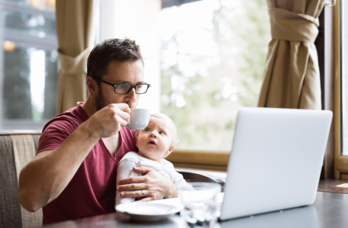 Ein Mann sitzt in einem Café, trinkt einen Kaffee, schaut auf einen Laptop und hält sein Baby fest.
