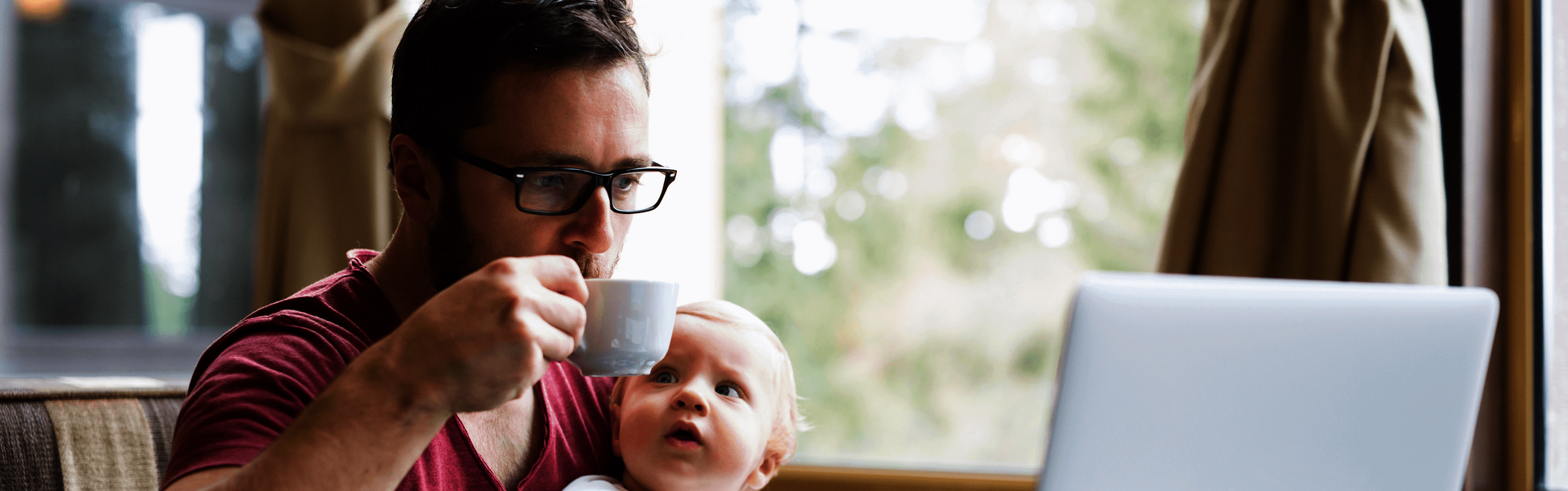 Ein Mann sitzt in einem Café, trinkt einen Kaffee, schaut auf einen Laptop und hält sein Baby fest.