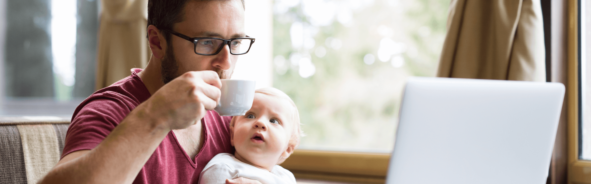Ein Mann sitzt in einem Café, trinkt einen Kaffee, schaut auf einen Laptop und hält sein Baby fest.