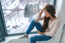 Ein Mädchen sitzt auf einer Fensterbank und schaut aus dem Fenster. Darußen ist es kalt und es liegt Schnee. Das Mädchen ist depremiert.